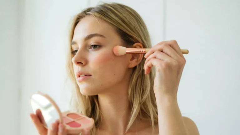 Aplicação de blush liquido com pincel nas maçãs do rosto: mulher aplica produto de maquiagem em frente a um espelho, com o cosmético rosado visível no estojo ao fundo.