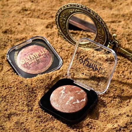 blush liquido em tons rosados com glitter, em embalagem compacta, sobre fundo de areia com acessórios ao redor.