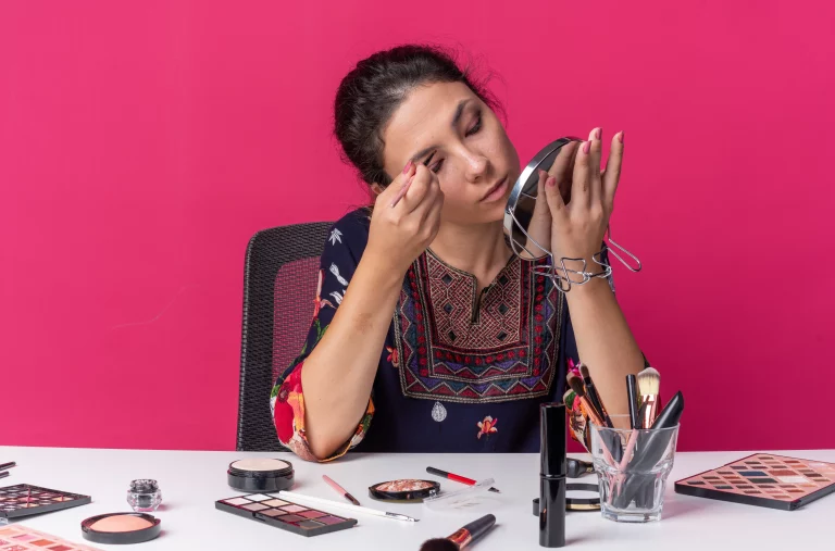 Mulher fazendo autoaquiagem com diversos produtos de beleza na mesa contra fundo rosa, focada na beleza e maquiagem.
