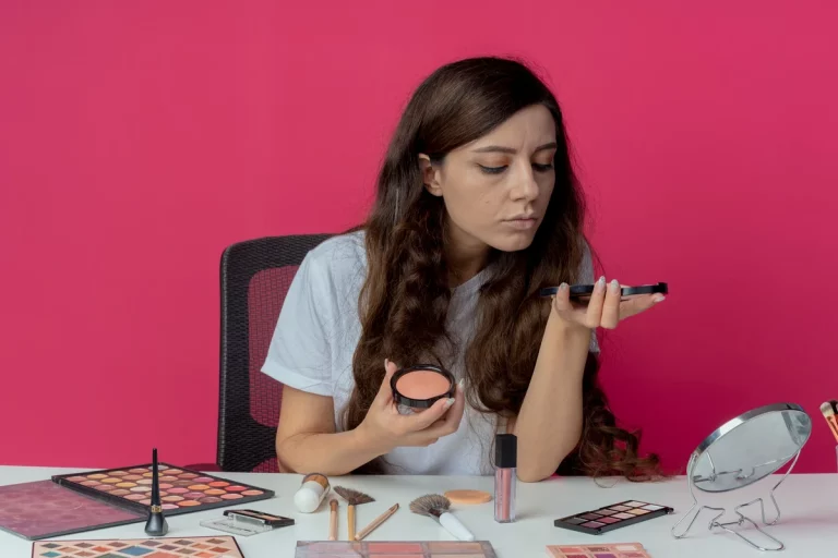 Uma mulher com cabelos longos e ondulados aplica maquiagem com um pincel, rodeada de produtos de maquiagem sobre a mesa, em um fundo rosa vibrante.