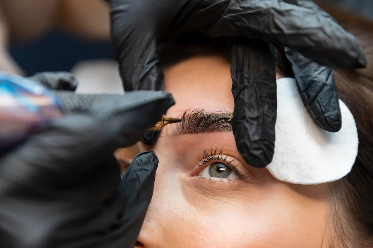 Pessoa com luvas pretas aplicando henna de sobrancelhas em uma mulher branca.