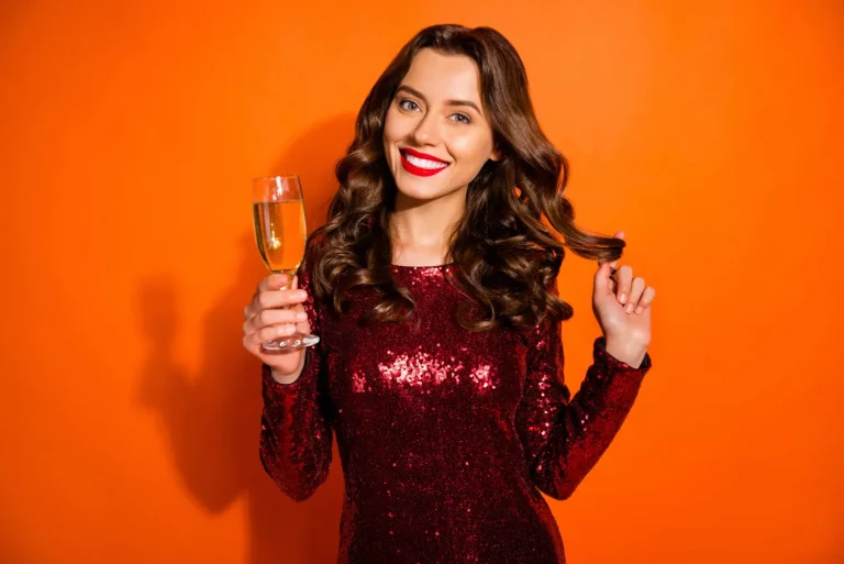 Mulher branca de vestido vermelho e com maquiagem para festa de final de ano, segurando taça e sorrindo.