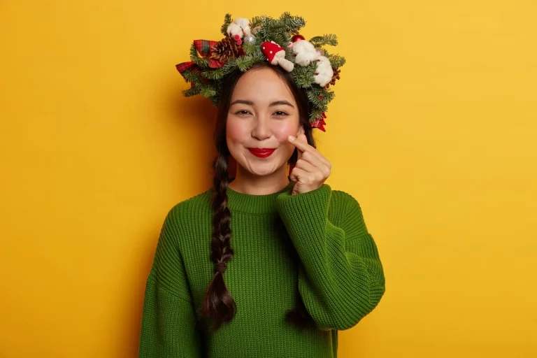 Mulher em fundo amarelo com acessórios natalinos sorrindo e fazendo símbolo de coração com os dedos
