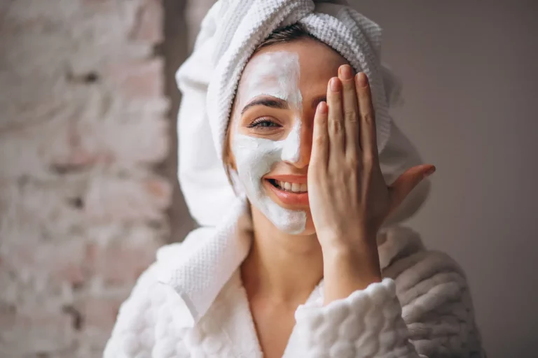 Mulher branca sorrindo com a mão cobrindo metade do rosto e na outra metade uma máscara em creme representando produtos skincare.