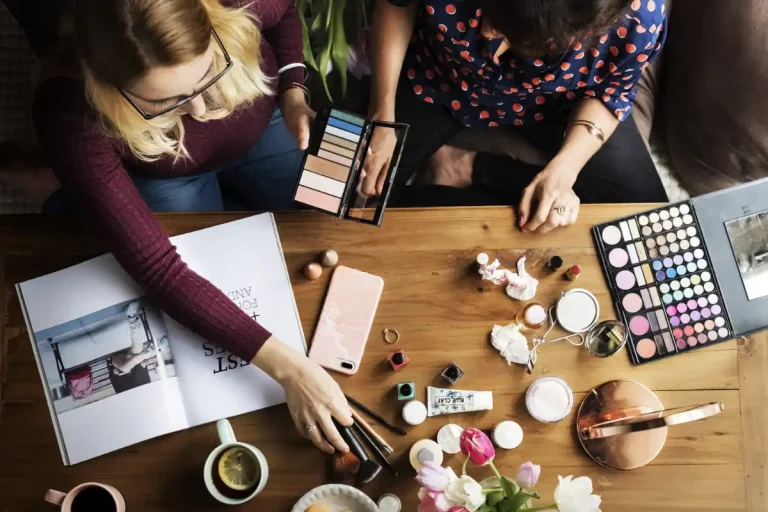 Duas mulheres aplicando maquiagem em uma mesa cheia de produtos.