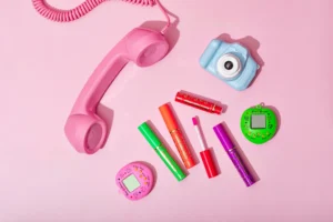 Coleção de batom mágico Bella Femme em cores vibrantes, incluindo telefone de brinquedo rosa, câmera de brinquedo azul, lançador de glitter e gadgets coloridos, tudo disposto sobre fundo rosa claro.