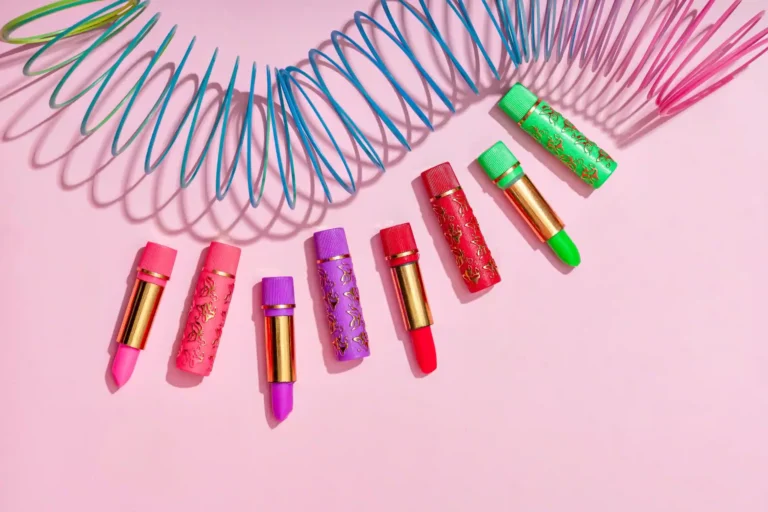 Batom mágico Bella Femme com embalagens decoradas em tons de rosa, roxo, vermelho, verde e azul, dispostos sobre fundo rosa claro ao lado de objetos coloridos.