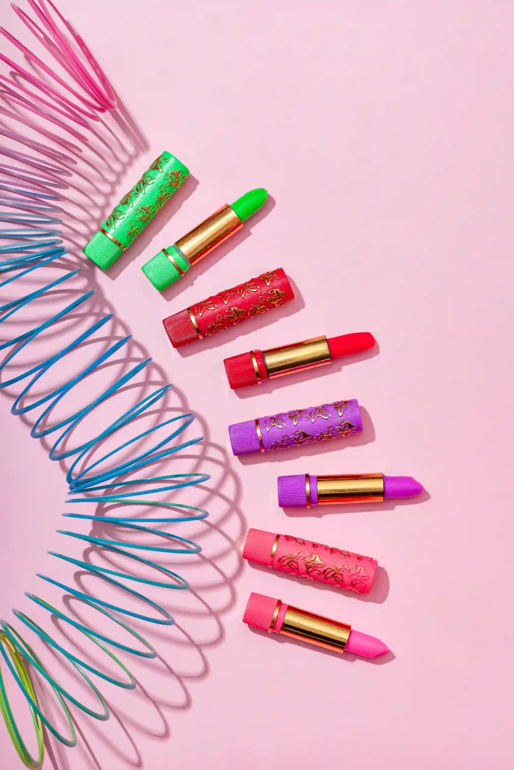 Batom mágico Bella Femme com embalagens decoradas em tons de rosa, roxo, vermelho, verde e azul, dispostos sobre fundo rosa claro ao lado de objetos coloridos.
