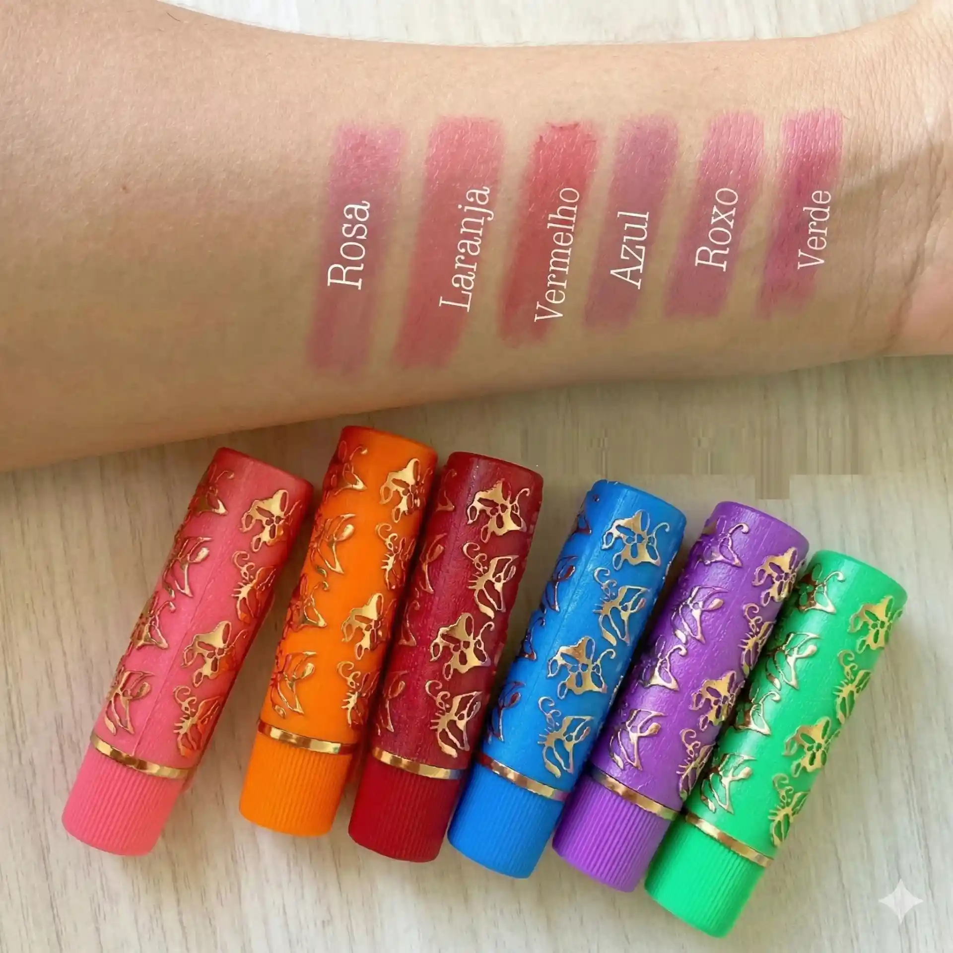 Cores de batom mágico Bella Femme em diferentes tonalidades, incluindo rosa, laranja, vermelho, azul, roxo e verde, demonstradas em um braço com swatches e batons decorativos na mesa.