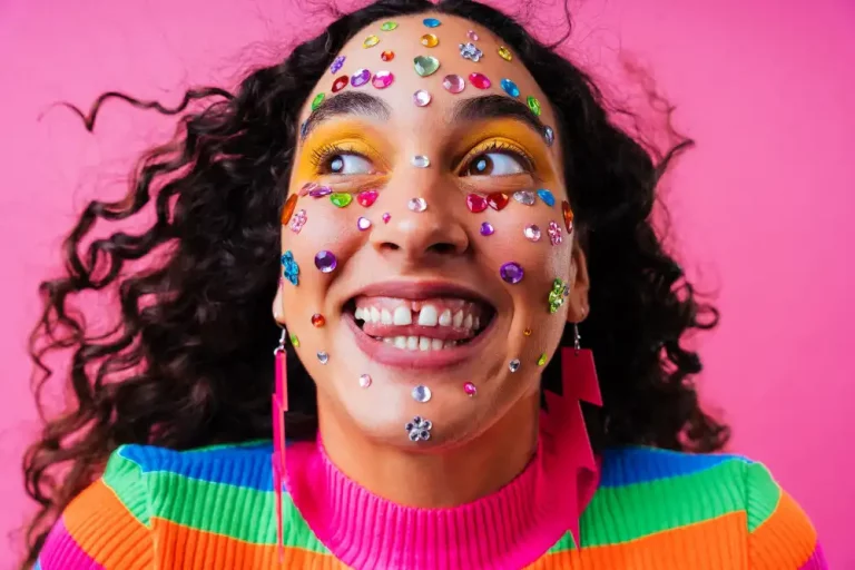 Mulher com maquiagem de carnaval simples, decorada com diversos adesivos coloridos no rosto, sorrindo e com expressão divertida, usando roupas vibrantes e fundo rosa.