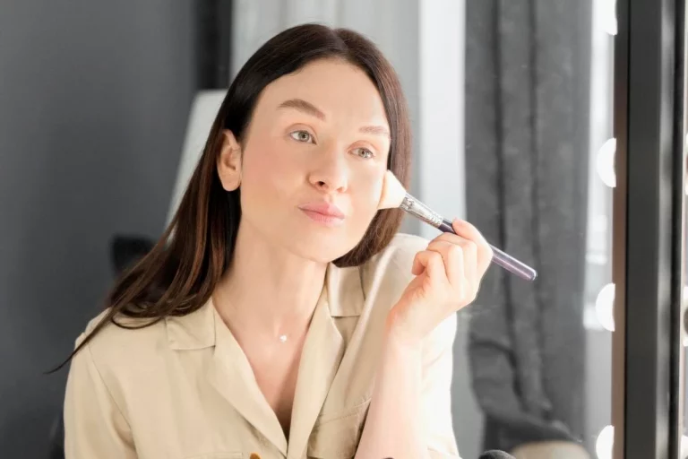 Mulher aplicando maquiagem no rosto com um pincel, refletindo em um espelho bem iluminado.