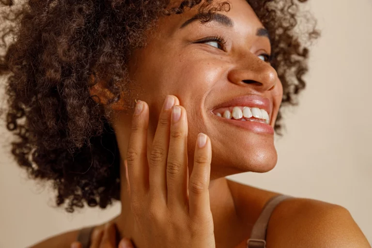 Close de uma mulher sorridente com cabelos cacheados, tocando suavemente o rosto, exibindo uma pele saudável e uma expressão alegre.