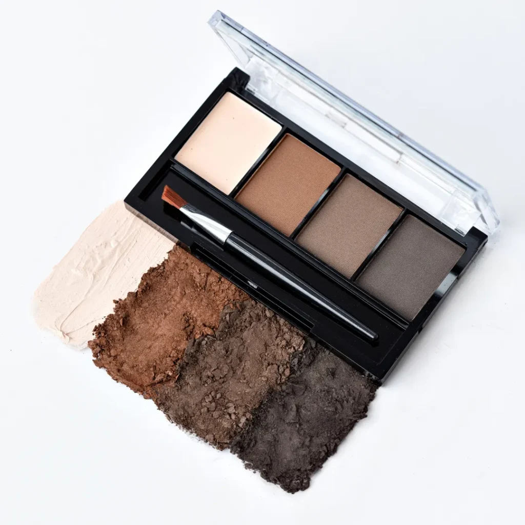 Estojo de Sombras e Primer para Sobrancelha com quatro tons de sombra em degradê, aberta sobre amostras de pó compacto claro, marrom e escuro em fundo branco, com pincel aplicador.