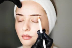 Aplicação de lápis dermatográfico no rosto: profissional com luvas usa lápis branco para marcar linha na sobrancelha e preparar o desenho durante procedimento estético, em close.