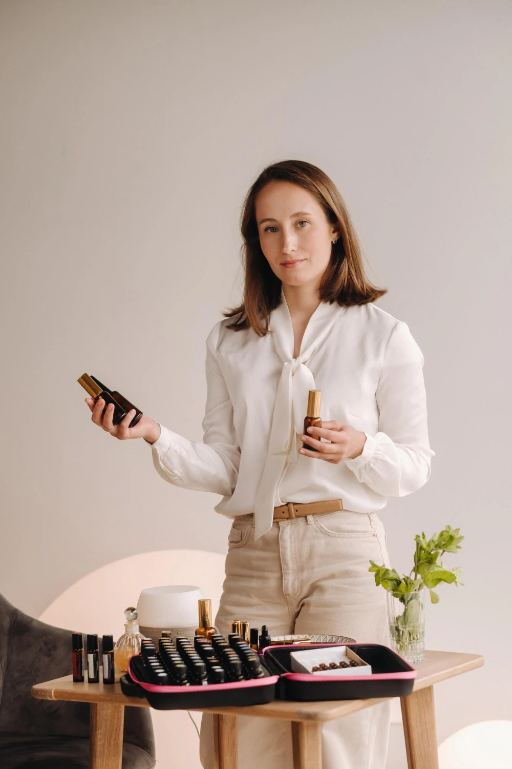 Mulher apresentando produtos de maquiagem em uma mesa com kits e frascos de cosméticos, segurando embalagem de base e iluminador; fundo neutro e clima profissional de beleza. fornecedor de maquiagem