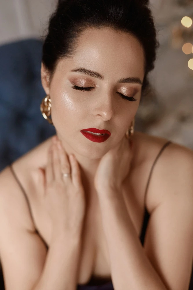 Mulher com maquiagem romântica para o dia dos namorados: olhos com sombra dourada suave e delineado esfumado, cílios destacados, e batom vermelho intenso em destaque, ao fundo com luzes desfocadas.