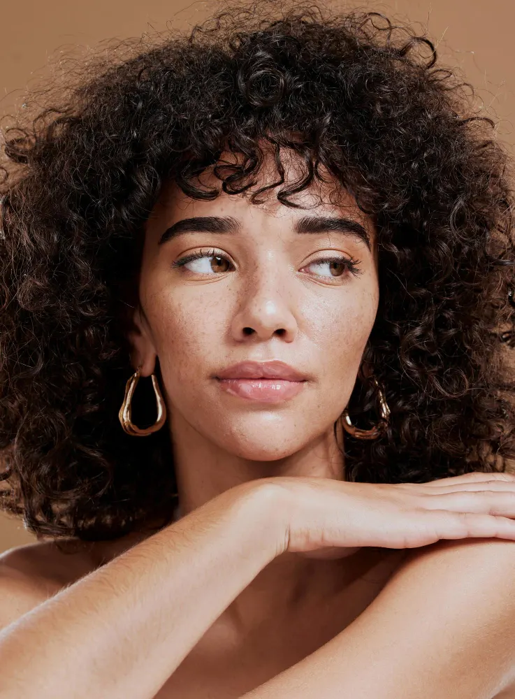 Mulher com maquiagem romântica para o dia dos namorados, com pele iluminada, sobrancelhas marcadas, batom rosado e expressão serena. Close no rosto com cabelo cacheado e fundo neutro.