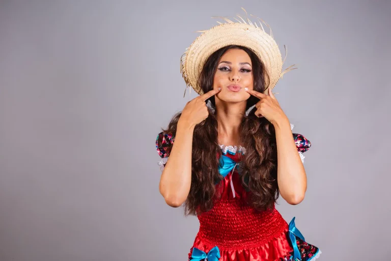 Mulher fazendo careta com dedos no rosto, maquiagem festa junina, usando chapéu de palha e vestido colorido, em cenário neutro, representando diversão e brincadeira.