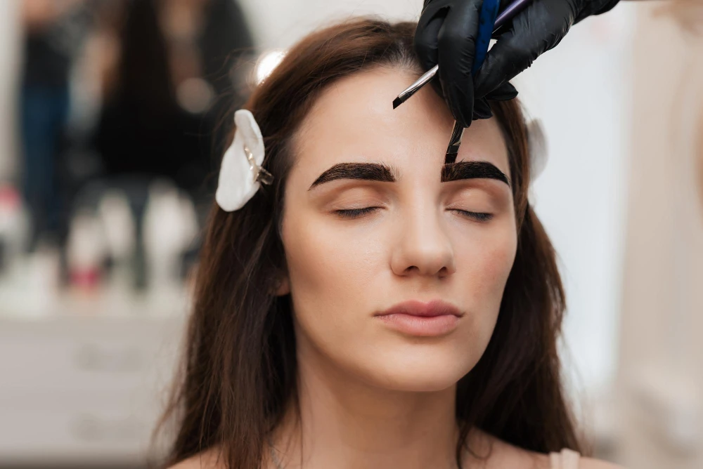 Aplicação de henna de sobrancelha em close: profissional com luva preta e pincel posicionando o produto sobre as sobrancelhas da cliente, com expressão relaxada.