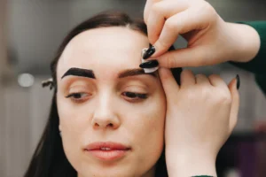 Aplicação de henna de sobrancelha: profissional desenha e preenche as sobrancelhas com pigmento usando pincel, enquanto a cliente mantém o rosto relaxado em ambiente de estética.