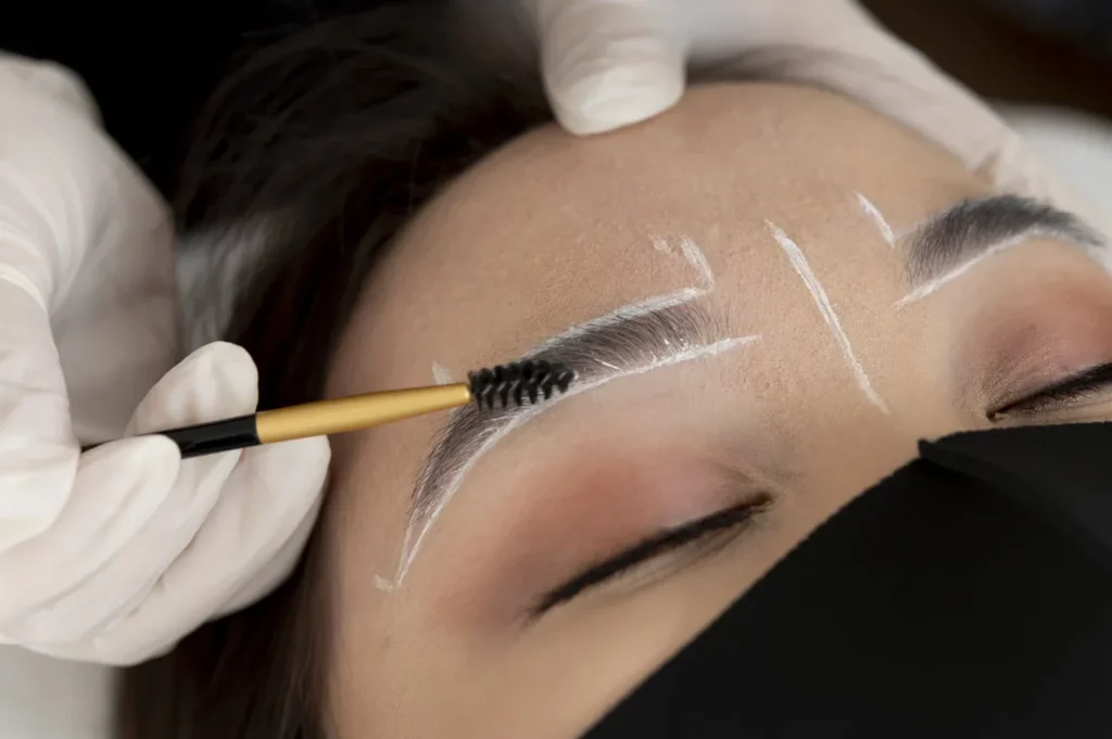 Profissional aplicando henna de sobrancelha com pincel em uma cliente, marcando o desenho das sobrancelhas sobre a pele com gel de cor e luvas brancas durante o procedimento