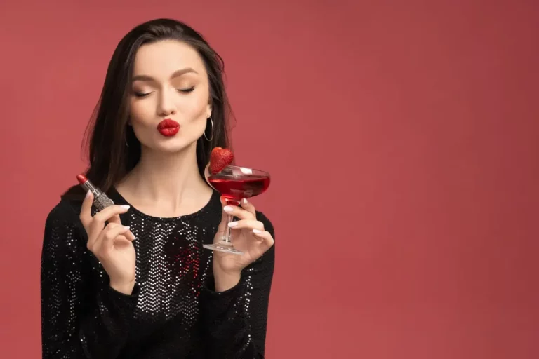 Mulher com vestido preto brilhante segurando batom matte vermelho e taça de coquetel com morango em fundo vermelho