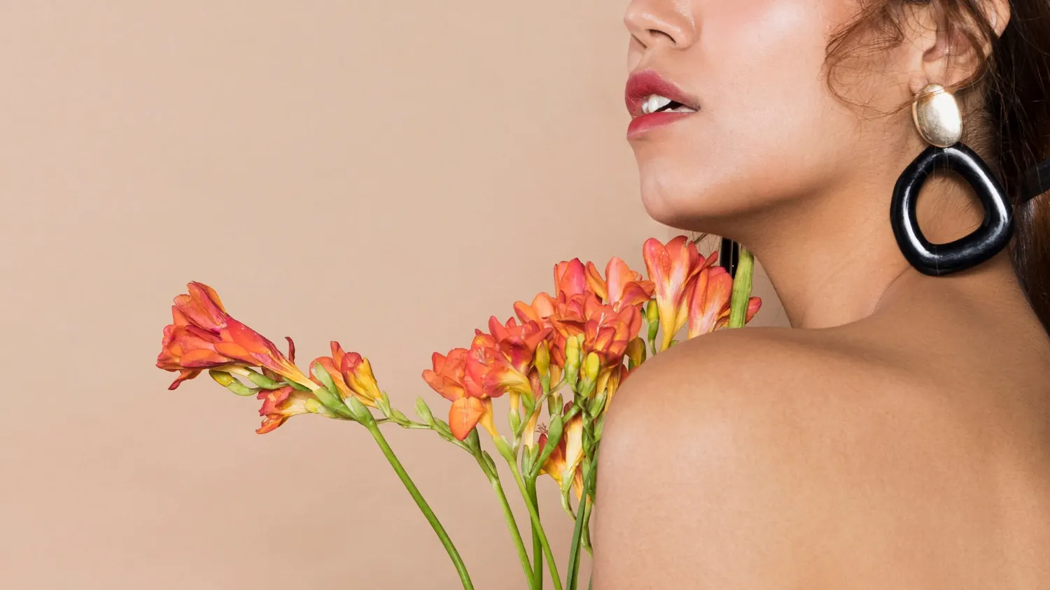 Mulher com brincos grandes e expressão serena, usando flores de cores vibrantes como cenário, com tons de laranja e rosa que remetem a cores da primavera em fundo claro e minimalista.