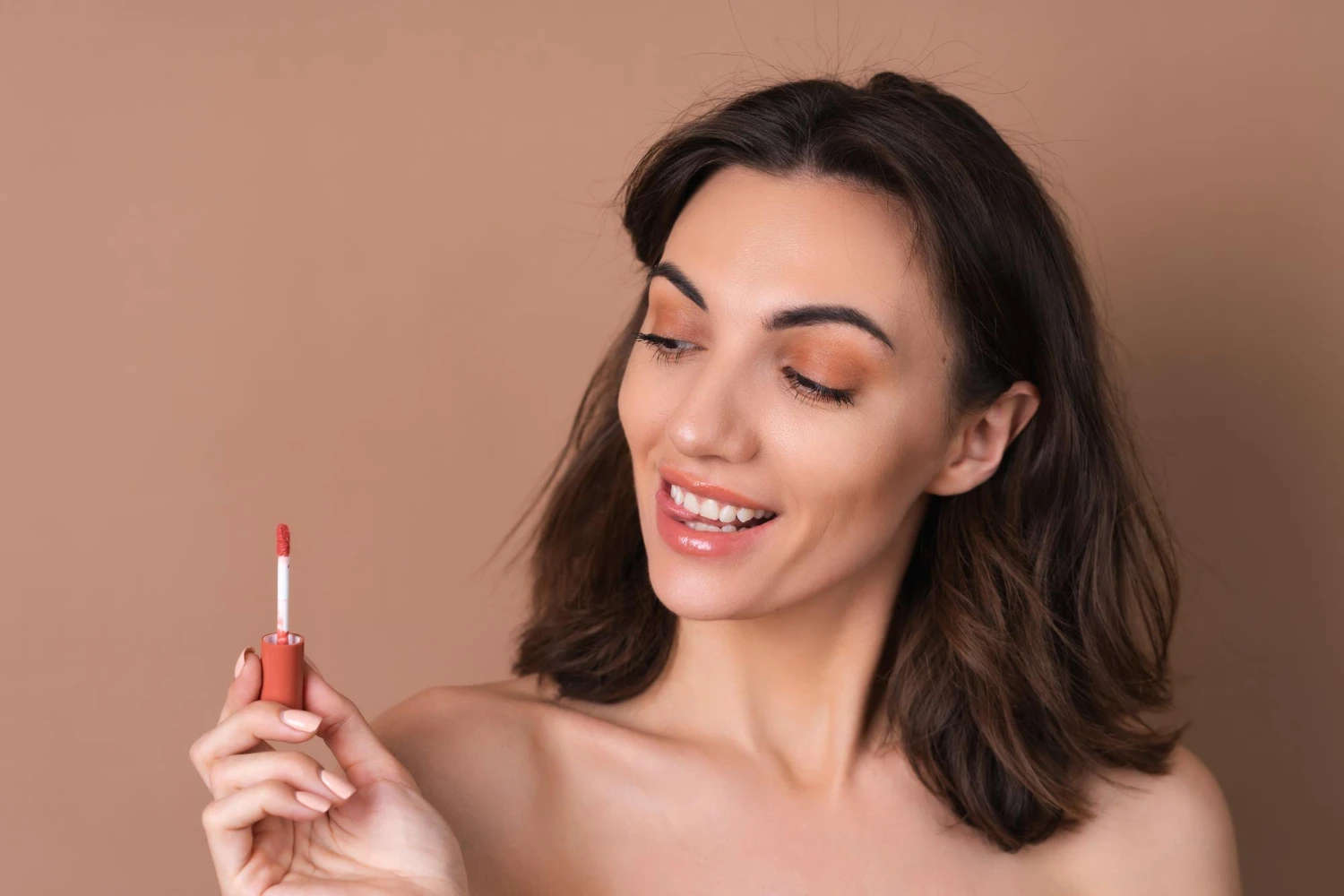 cores da primavera: Mulher com cabelo escuro e maquiagem suave sorrindo enquanto segura um batom vermelho, com fundo neutro e iluminação suave, destacando cores vivas e primaveris