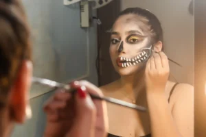 Maquiagem Halloween com maquiagem de caveira no rosto, glitter dourado nos olhos e acabamento branco nos dentes, enquanto a pessoa aplica o efeito no espelho.