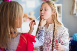 Criança recebendo maquiagem infantil na sala, enquanto uma pessoa aplica maquiagem com pincel e a menina segura um batom, com roupas brilhantes em clima de faz de conta.