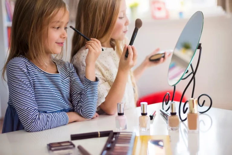Duas crianças se maquiando e brincando de maquiagem infantil em frente a um espelho, com diversos cosméticos Maria Pink e pincéis sobre uma mesa.