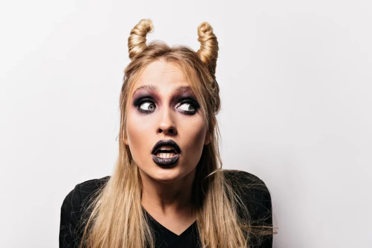 Mulher com maquiagem de halloween dramática, cabelo com dois coques e expressão de surpresa, usando roupas pretas para o tema gótico ou alternativo.
