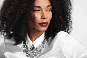 Retrato de mulher de pele negra com maquiagem natural e iluminada, cabelo cacheado e colar de pedrarias, destacando acabamento de pele e lábios em tom marrom.