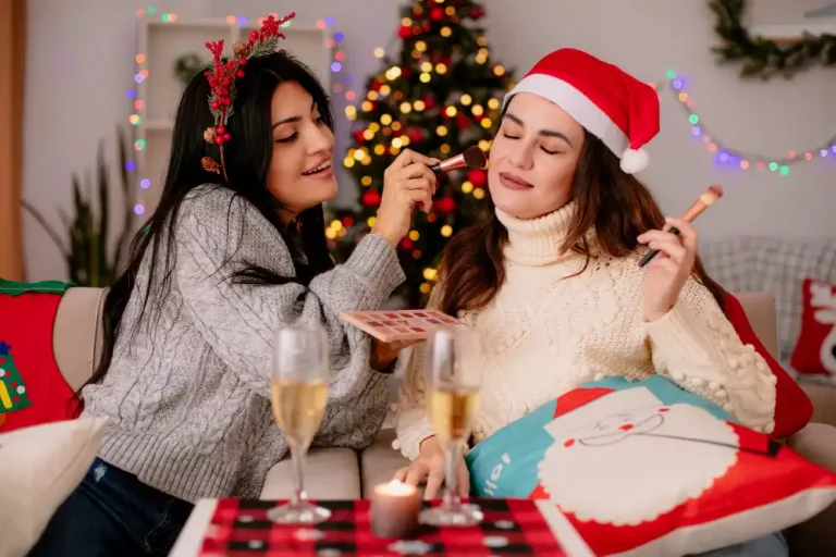 Duas mulheres comemorando o Natal, uma delas recebendo maquiagem enquanto a outra se diverte, com decoração natalina ao fundo. Revenda de maquiagem para o natal
