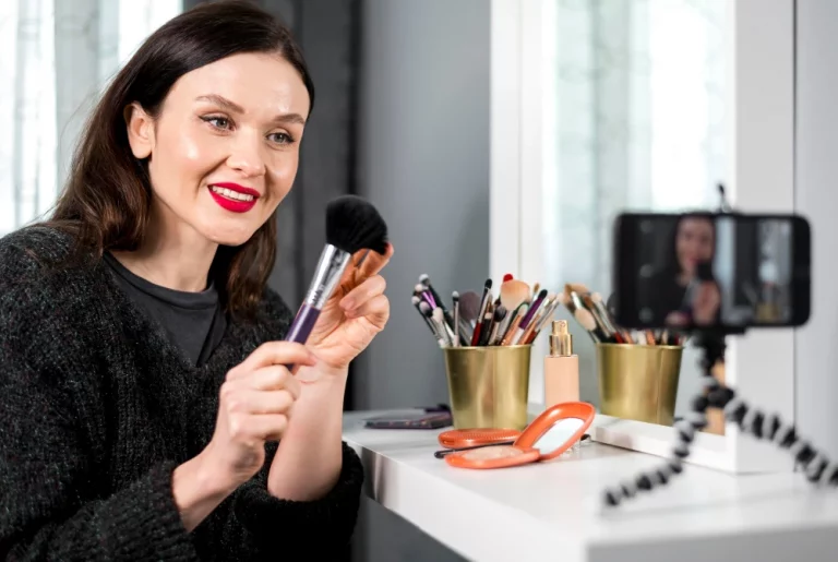 Mulher criativa gravando vídeo sobre estratégias de vendas, usando maquiagem e produtos de beleza, em ambiente bem iluminado, com celular em suporte e decoração moderna.