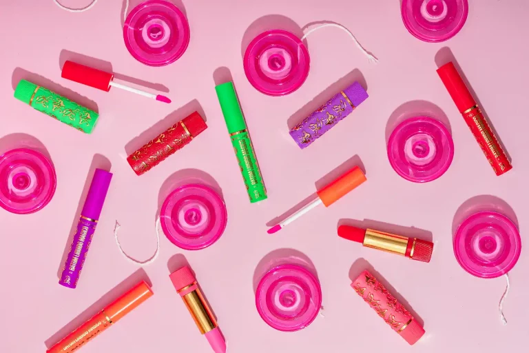 Diversos produtos de maquiagem e cosméticos em tons de rosa, roxo, verde e vermelho, dispostos em um fundo rosa claro, ilustrando o guia completo Bella Femme.