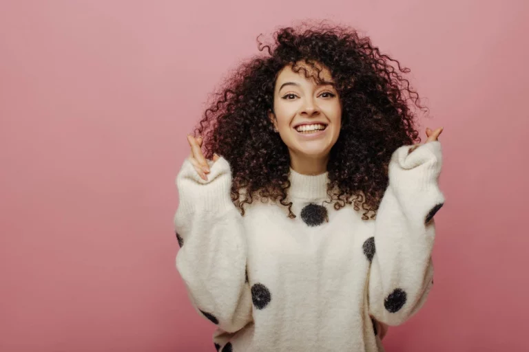 Mulher feliz com cabelo cacheado, vestindo moletom branco com pontos pretos, sorrindo alegremente em fundo rosa, transmitindo felicidade e positividade.