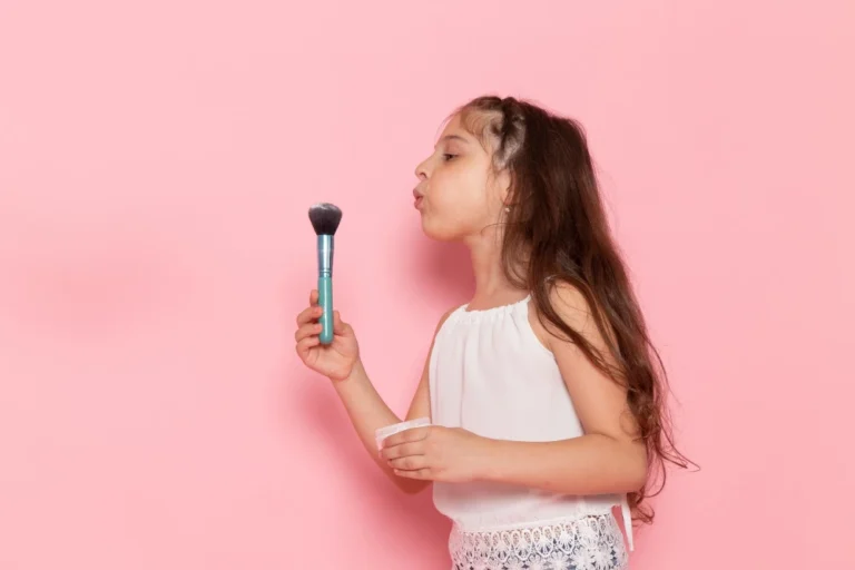 Menina com longos cabelos castanhos segurando um pincel de maquiagem com os olhos fechados em um fundo rosa. Conteúdo sobre marcas de cosméticos para revender, apresentando a Maria Pink da Bella Femme