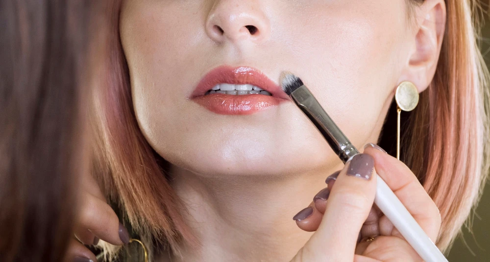 Mulher recebendo maquiagem para noiva nos lábios com um pincel, destacando o processo de maquiagem e beleza, com foco na preparação para um evento ou sessão de fotos.