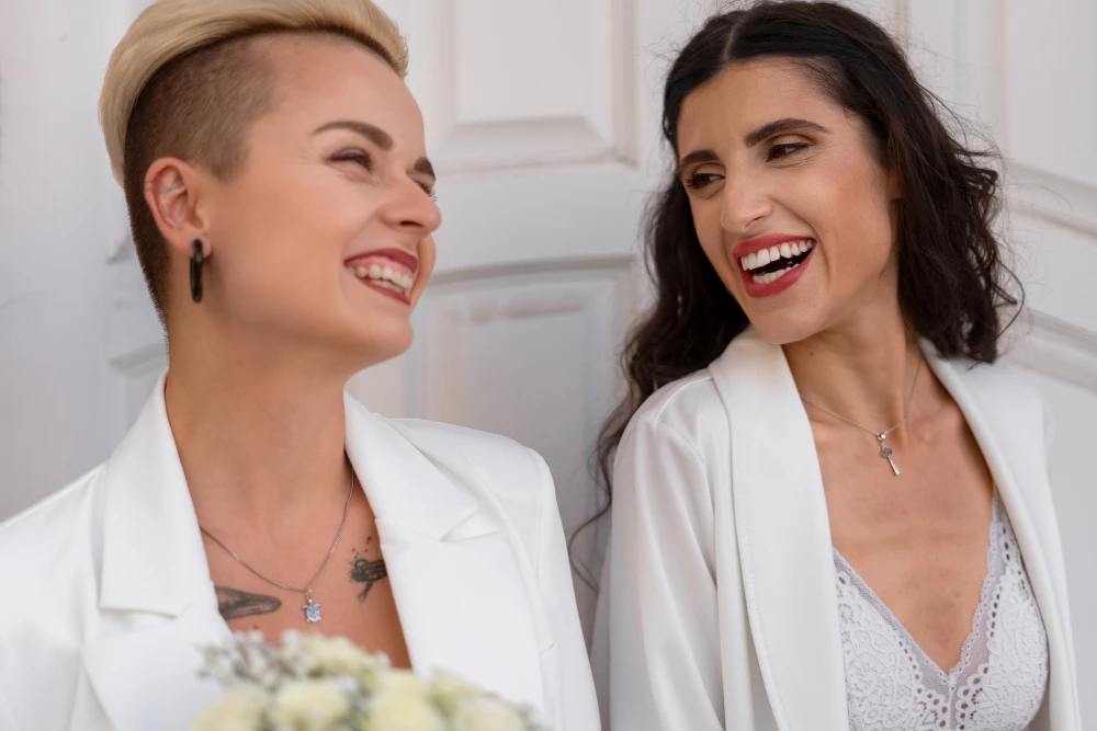 Mulher com cabelo com estilo undercut e outra com cabelo cacheado, ambas casando, sorrindo e usando roupas brancas, em um momento de alegria. Conteúdo sobre maquiagem para noiva.