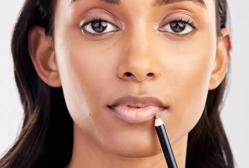 Close no rosto de uma mulher aplicando maquiagem nos lábios com um batom labial, mostrando um lip combo com acabamento natural e iluminado em pele clara contra fundo claro.