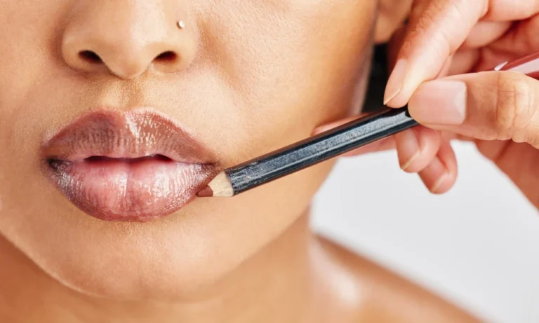 Close-up de uma mulher aplicando batom com um aplicador fino em estilo lip combo, com a boca e o lábio bem destacados e mão segurando o cosmético sobre fundo claro