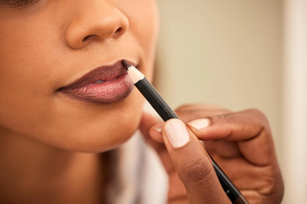 Close-up de uma aplicação de lip combo: mãos usando um pincel aplicador com lápis de lábios para contornar e preencher a boca com batom em tons rosados e marrons, com pele e acabamento bem definido.