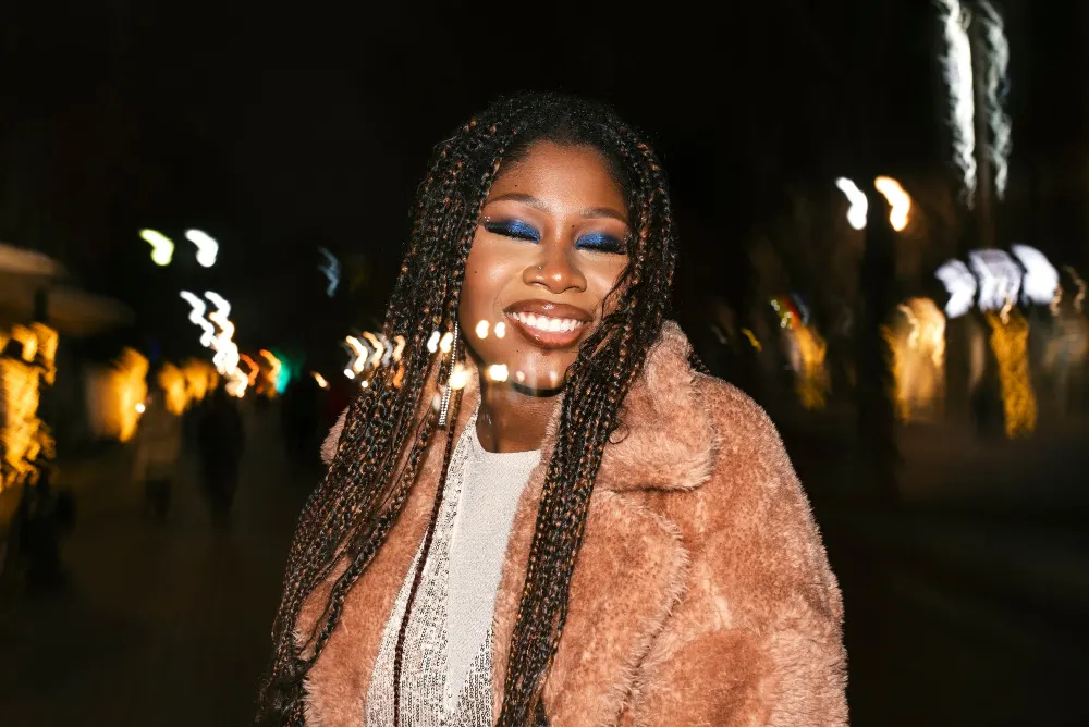 Mulher sorridente com tranças e maquiagem marcante, usando casaco de inverno marrom com acabamento em pele, em ambiente externo à noite com iluminação desfocada ao fundo, destacando tendências de inverno.
