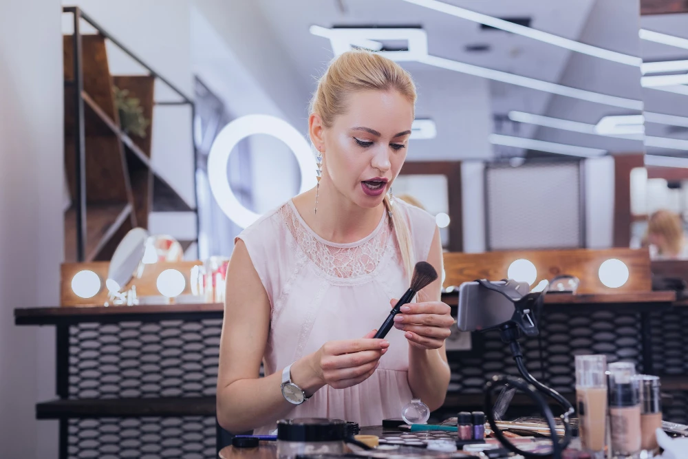 Maquiagem em ambiente de estúdio: mulher aplica maquiagem com pincel e espelho portátil, com produtos e luzes ao fundo, ideia de tendências de inverno e beleza