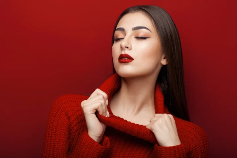 Modelo com suéter vermelho de tricô e maquiagem elegante, com expressão serena e fundo vermelho liso, destacando tendências de inverno em moda e beleza.