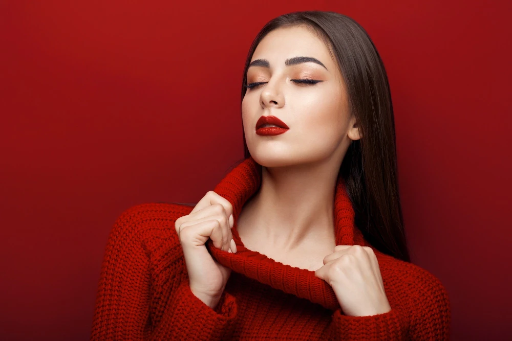 Modelo com suéter vermelho de tricô e maquiagem elegante, com expressão serena e fundo vermelho liso, destacando tendências de inverno em moda e beleza.