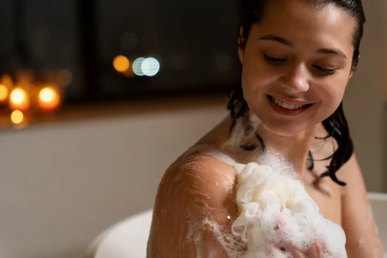 Mulher no banho premium, com espuma e água escorrendo sobre o ombro e o corpo, sorrindo enquanto relaxa em um ambiente acolhedor com luzes ao fundo.