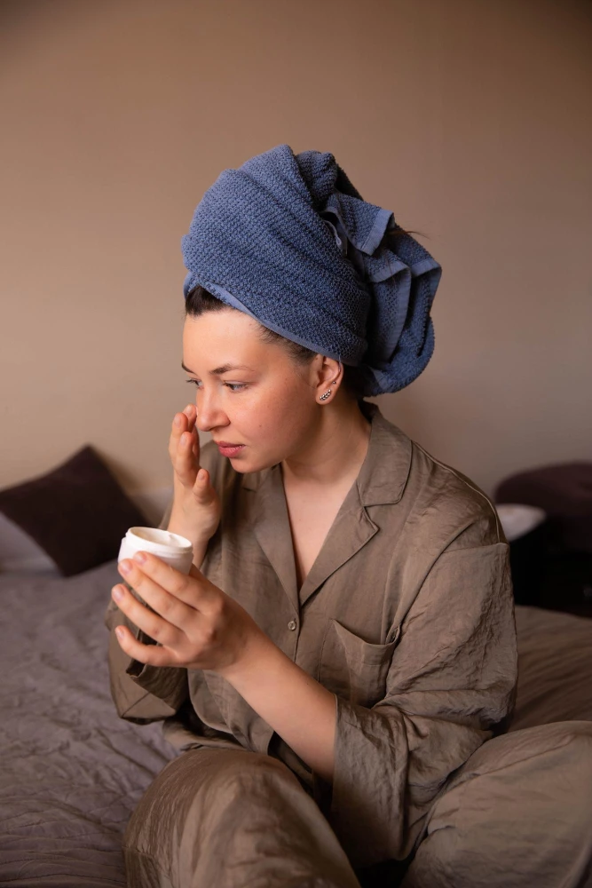 Mulher com touca turquesa e pijama em casa, aplicando creme hidratante com os dedos no rosto. Cena iluminada em ambiente aconchegante, sugerindo cuidados com pele no inverno.