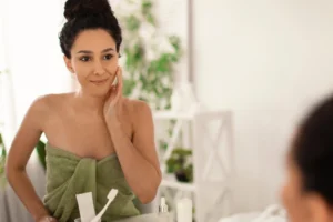 Mulher com toalha no cabelo e rosto hidratado em frente ao espelho, aplicando produto facial com a mão; ideal para rotina de cuidados com pele mista em casa.
